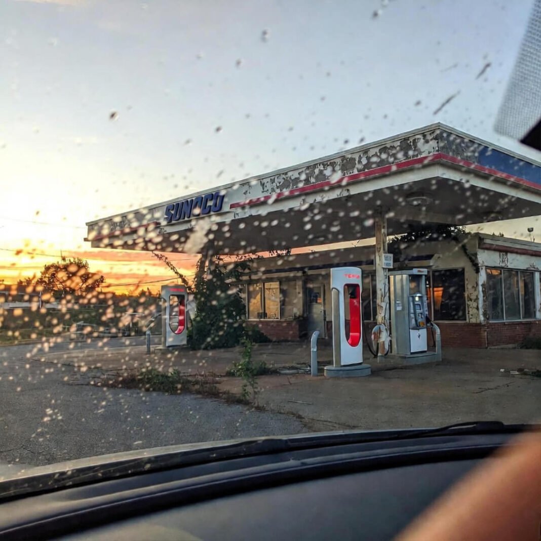 A vintage-inspired digital illustration of a person hunched over the manual shifter of a Ford Mustang EV prototype in a dimly lit garage. A crumpled blueprint lies on the seat.